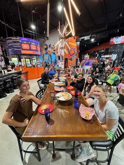 Friends gathered around a long table in a colorful arcade, smiling and eating together.