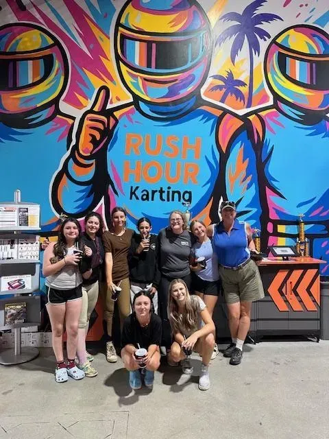 Group posing in front of colorful Rush Hour Karting mural and kart display, making celebratory hand gestures.