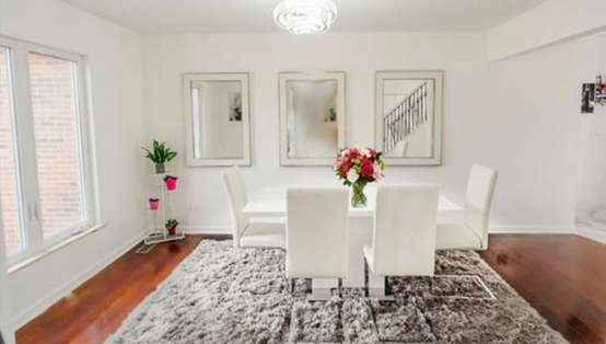 A white dining room with table, chairs, rug, and three mirrors above it. Wooden floor, and a small plant stand by the window.