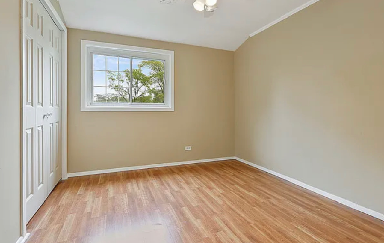 Empty bedroom with tan walls, wooden floor, white door and window; daylight shines through the window.