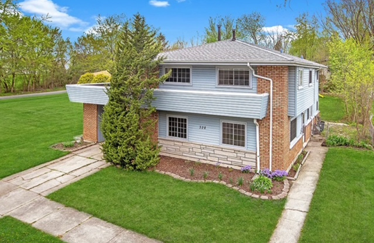 Two-story house with light blue siding, red brick accents, and a concrete driveway, set on a green lawn with trees.