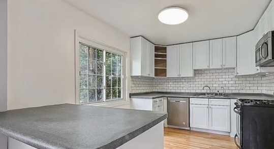 White kitchen with a gray countertop, stainless steel appliances, and a window with a view of greenery.