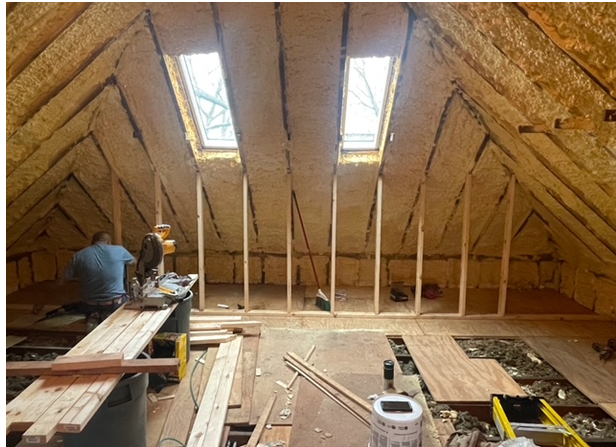 Interior view of an attic under renovation, with insulation between rafters. A worker is cutting wood.