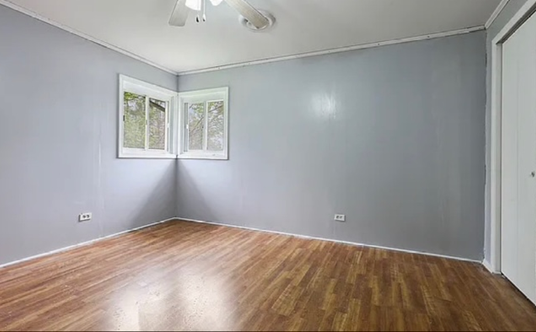 An empty room with hardwood floors , gray walls and two windows.