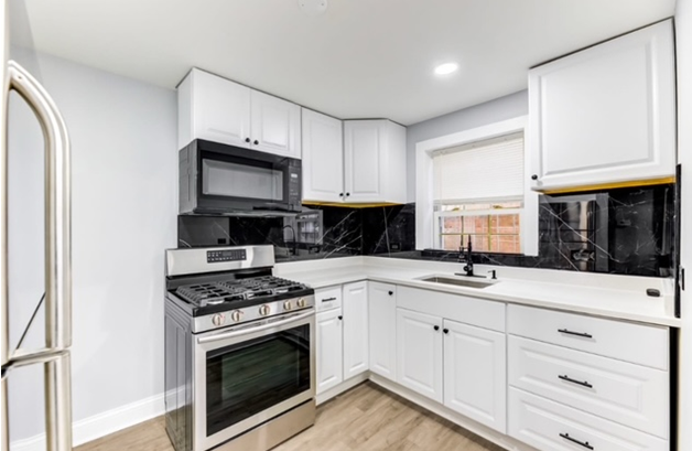 White kitchen with stainless steel appliances, including stove and microwave. Black marble backsplash and light wood floors.