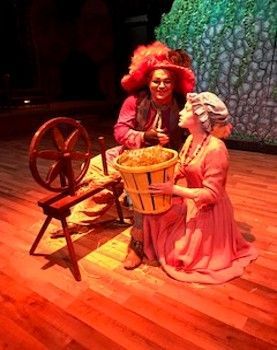 Two costumed performers kneel by a wooden spinning wheel and basket on a stage with warm lighting
