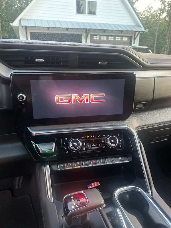 GMC truck dashboard with center display showing GMC logo, parked in a driveway with a house ahead