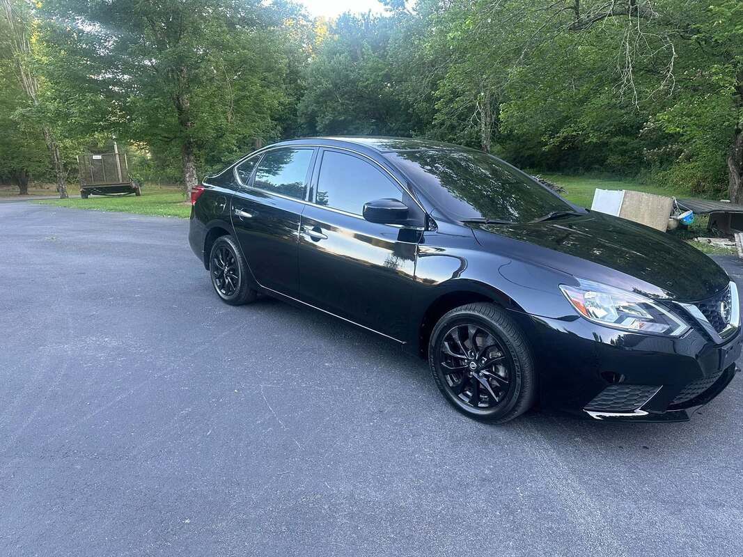 Black sedan parked on a paved lot beside trees, with a bench in the background.