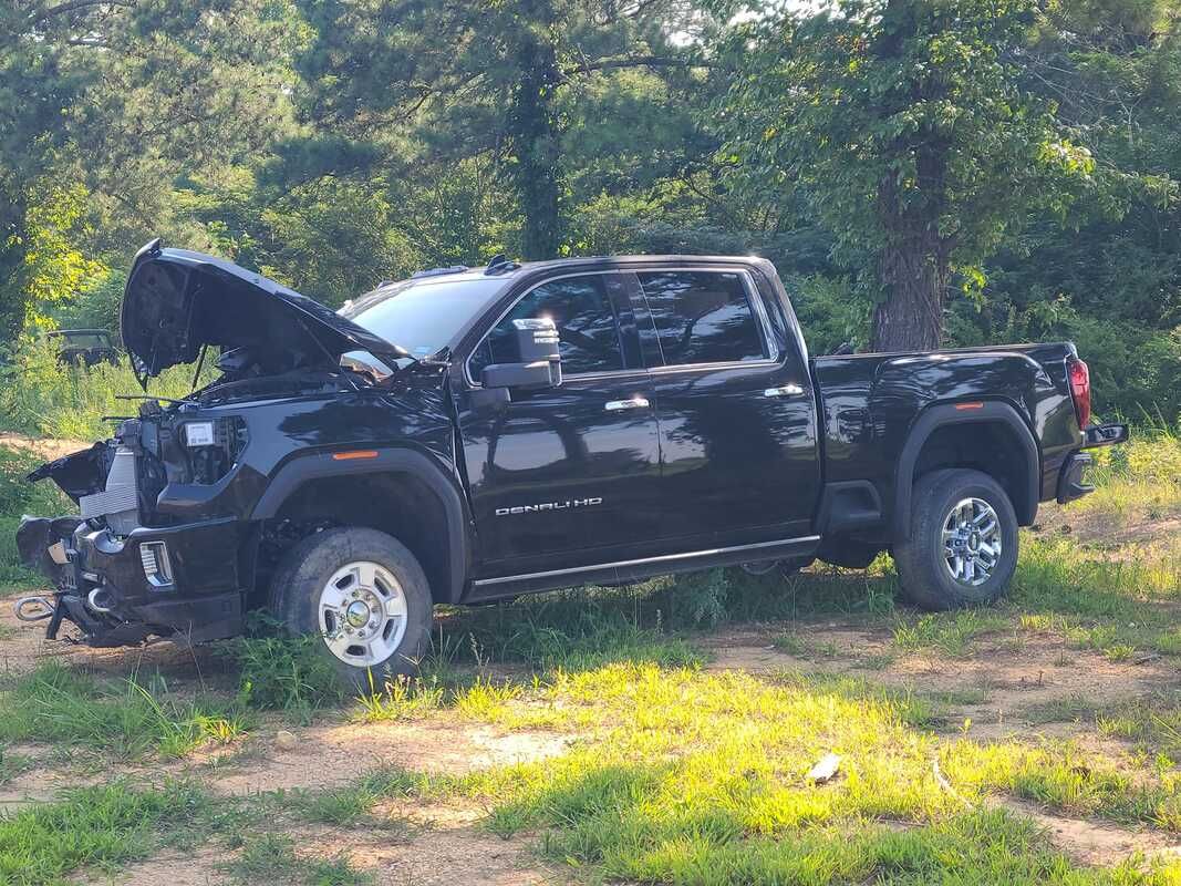 Black pickup truck with front end damage and hood open, parked on grass by trees.