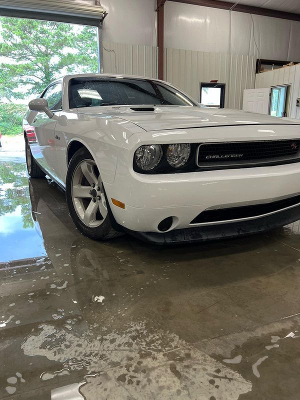 White Dodge Challenger in a garage, front-left view, with wet concrete floor and open door to daylight