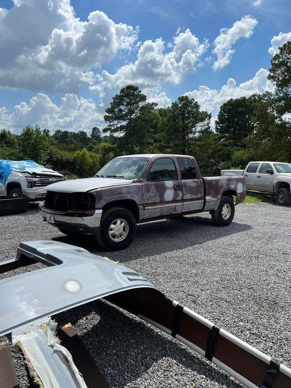Old pickup truck parked on gravel in a lot under a cloudy blue sky, with other vehicles nearby