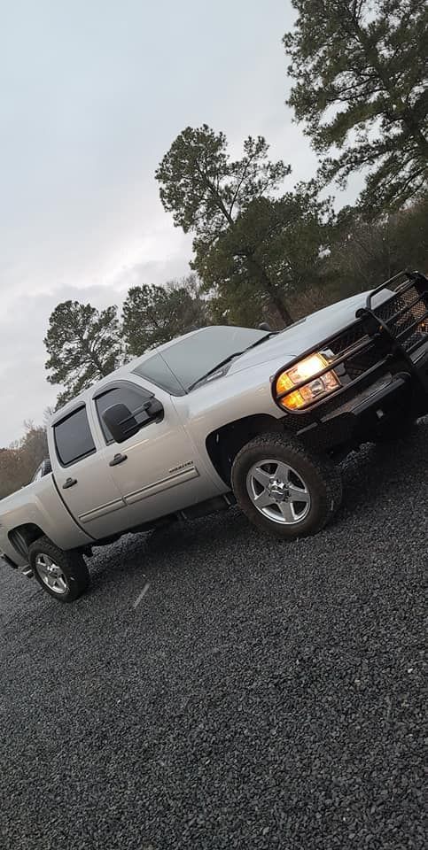Silver pickup truck parked on gravel road, angled front view with headlights on near trees.