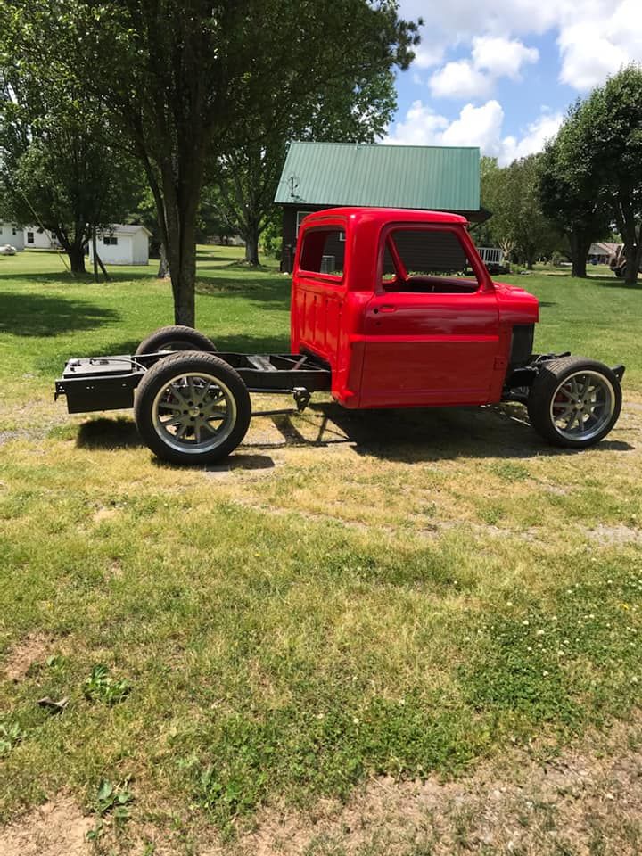 Red custom truck chassis with bright red cab parked on grass under trees near a small building.