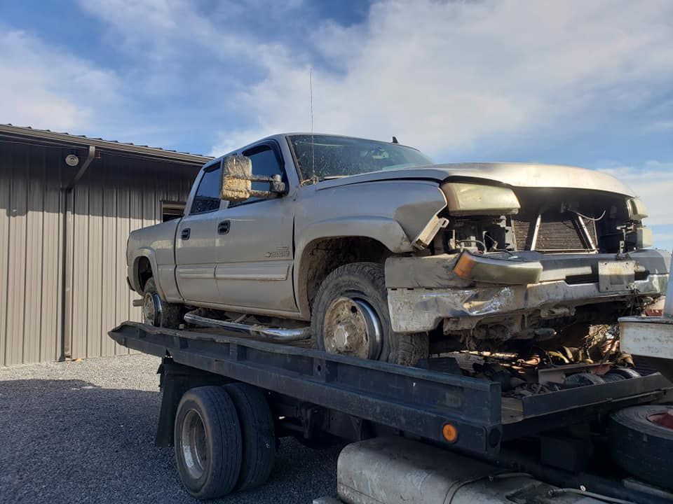 Damaged silver pickup truck on a flatbed trailer outside a garage, front end heavily crushed.