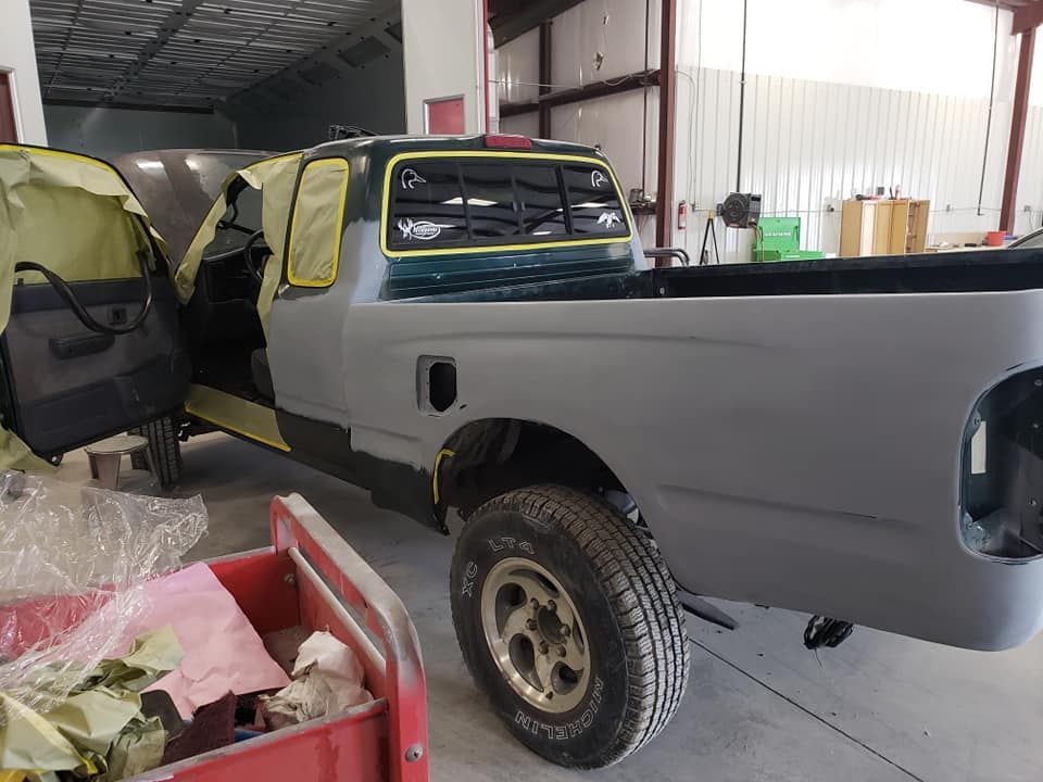 Gray pickup truck in a garage with taped windows and open door, amid auto repair supplies.