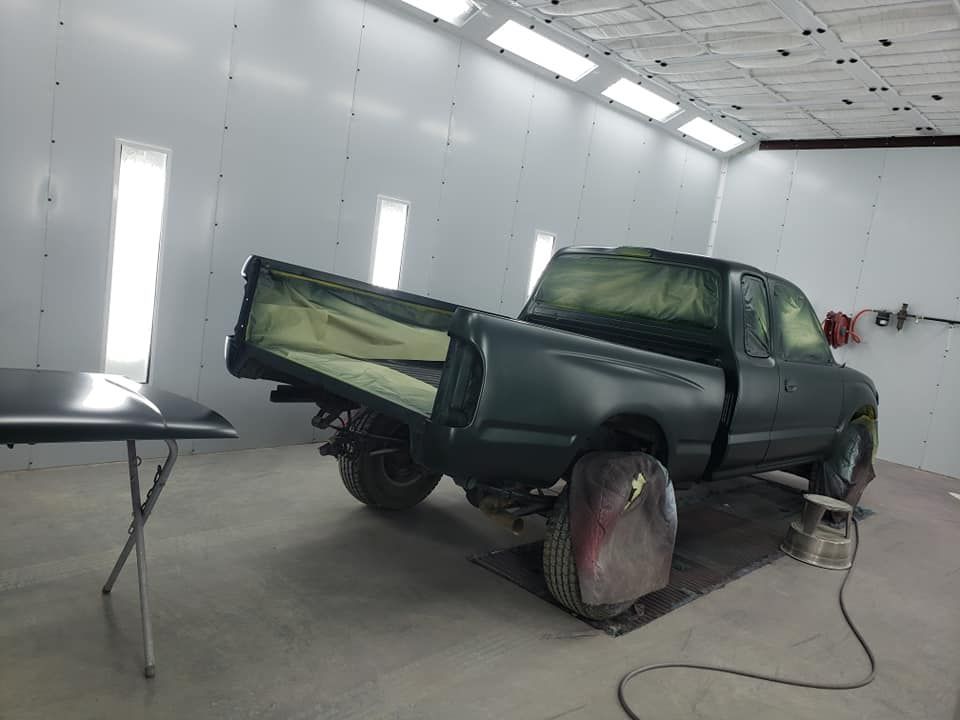 Green pickup truck in a paint booth, partly disassembled with rear wheel removed.