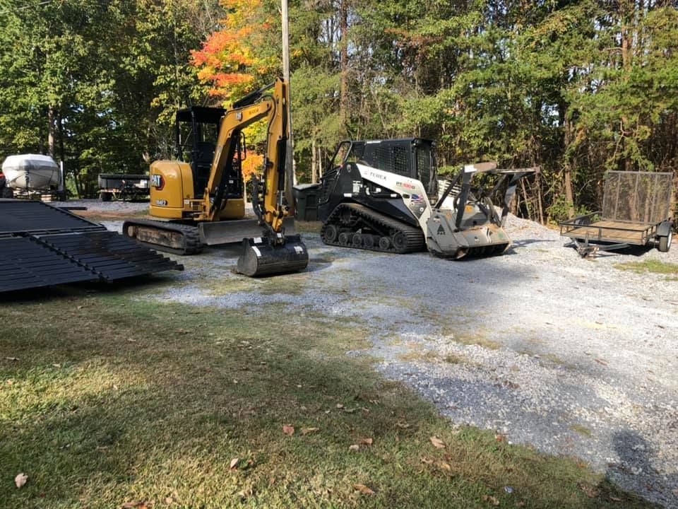 Yellow excavator and white skid steer on a gravel lot beside a wooded area.