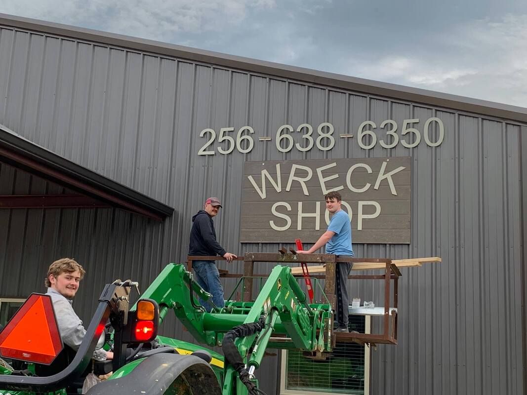 People on a green tractor lift a sign reading “NREP SHOP” on a gray metal building.