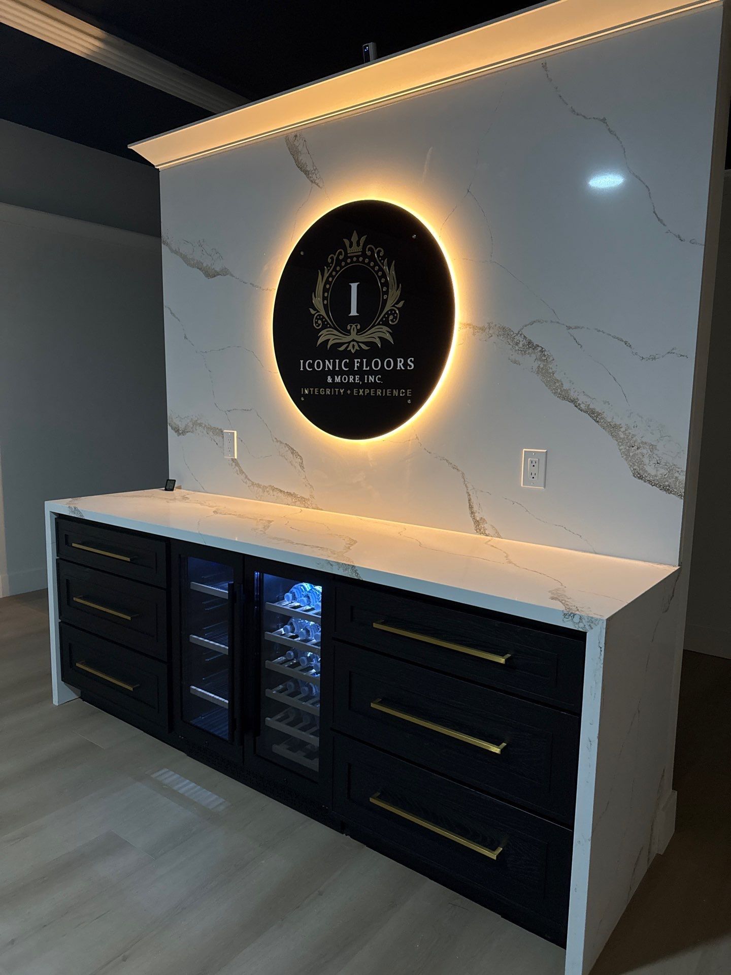 Bar area with black cabinets, white countertop, wine fridge, and lit logo on marble wall.