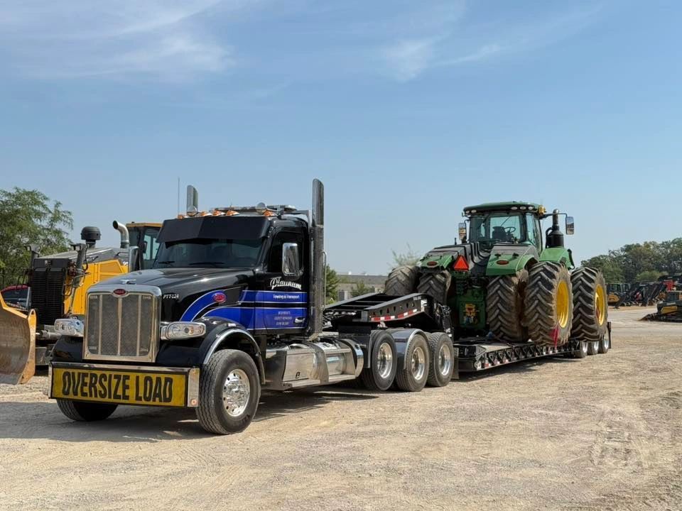 Black semi-truck hauling a green John Deere tractor on a trailer, labeled 