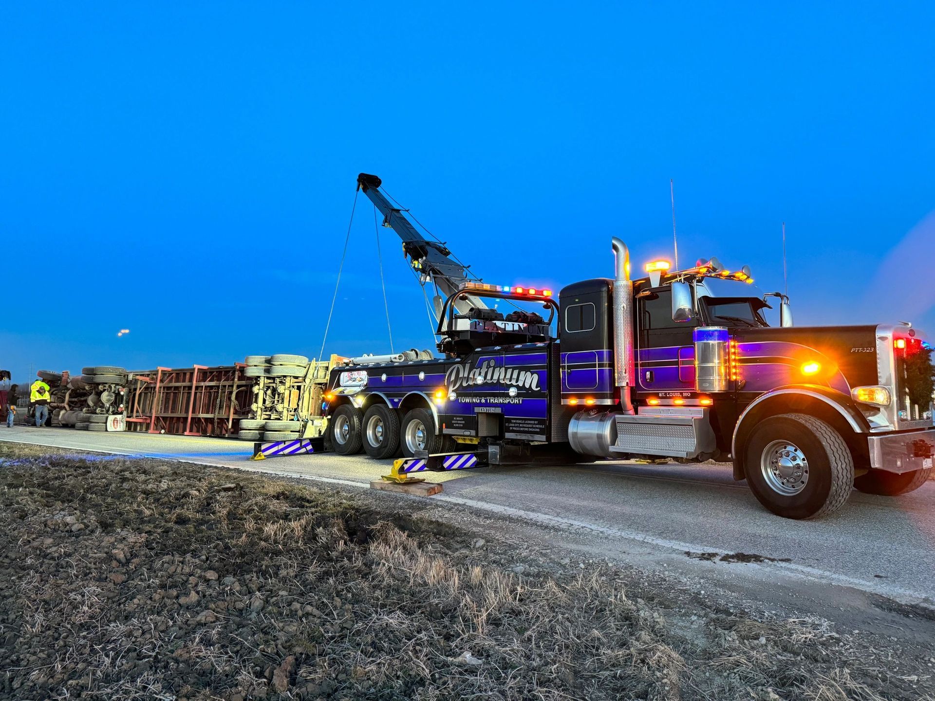 Tow truck uprighting a semi-trailer on a roadside under a blue dusk sky.