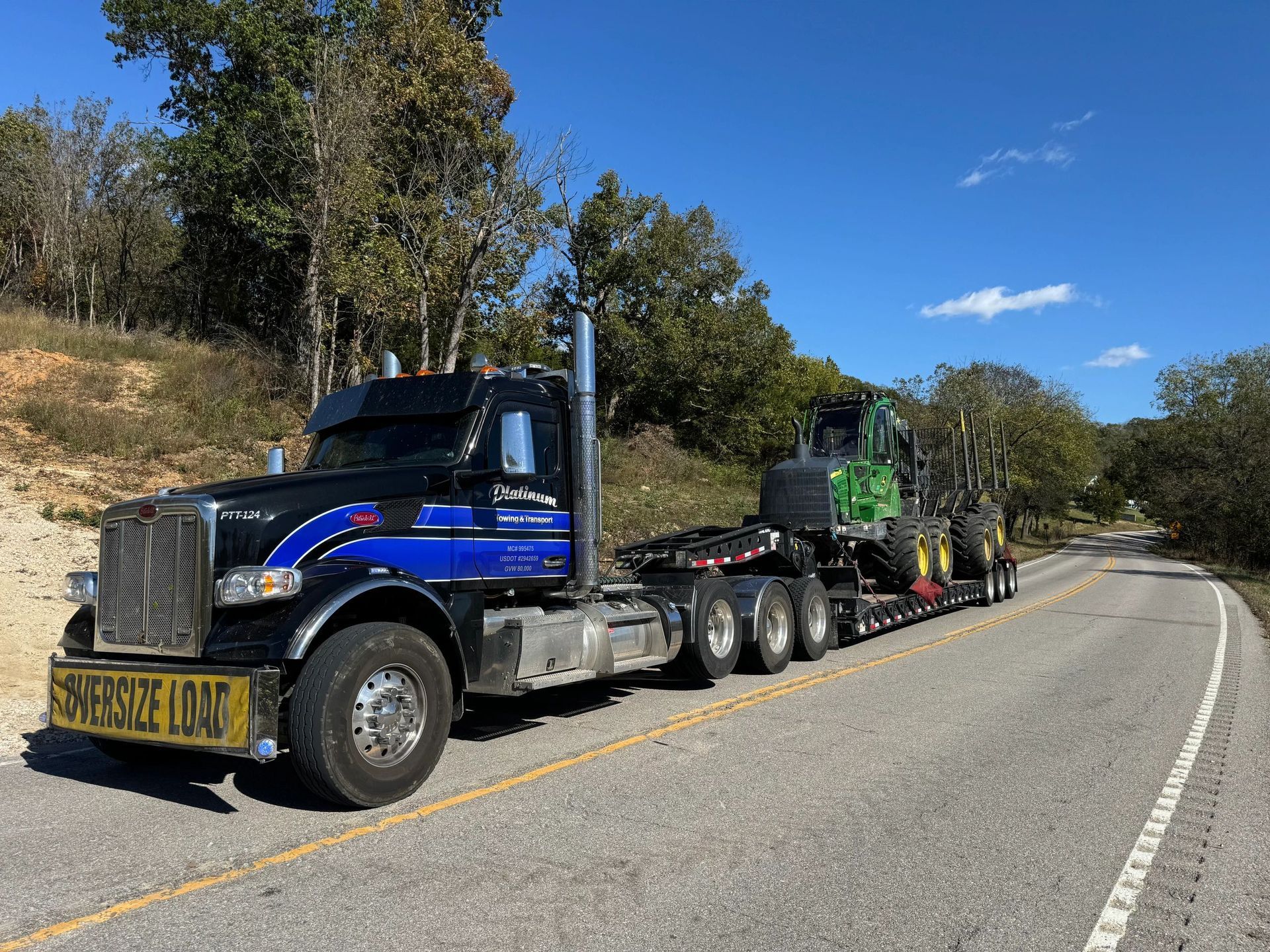 Black semi-truck hauling green forestry equipment on a curved road, under a blue sky. 