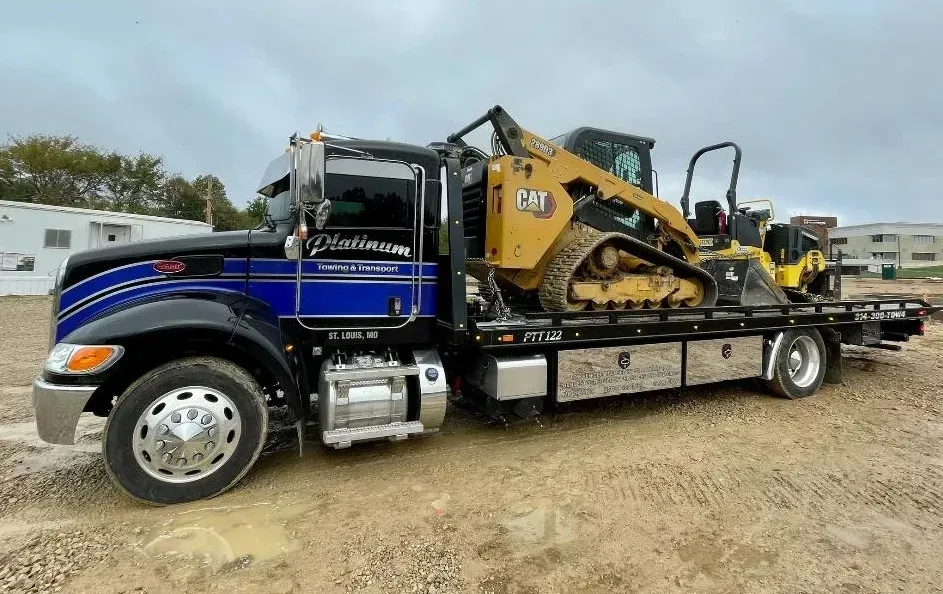 Flatbed truck carrying a yellow Caterpillar skid steer on a construction site.