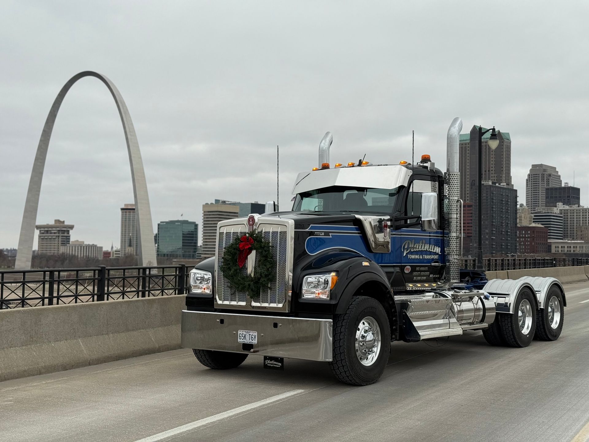 Semi-truck with a wreath on the front, driving on a bridge in front of the Gateway Arch and St. Louis skyline.