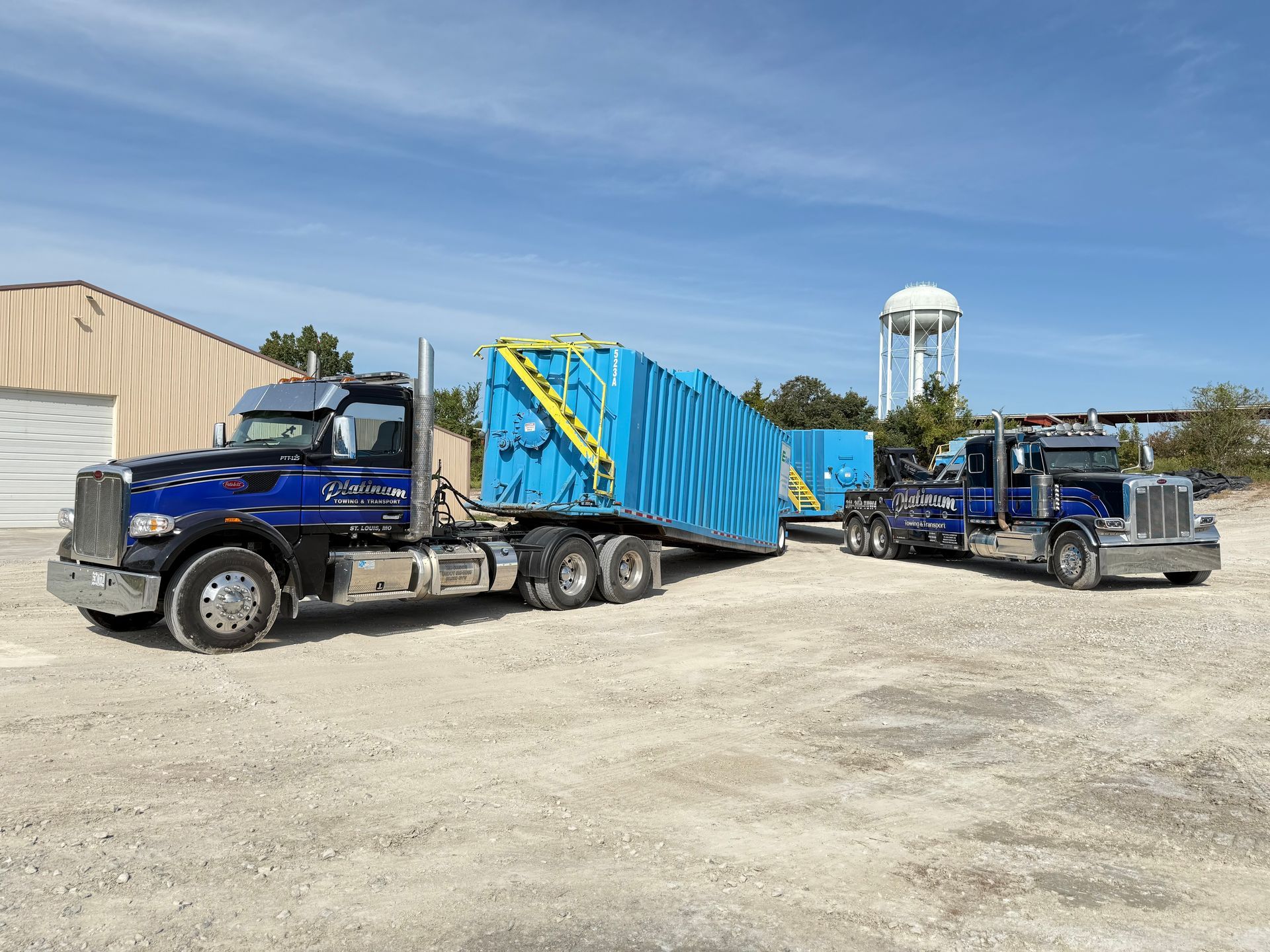 Two blue semi-trucks transporting large blue rectangular objects on a gravel lot under a blue sky.
