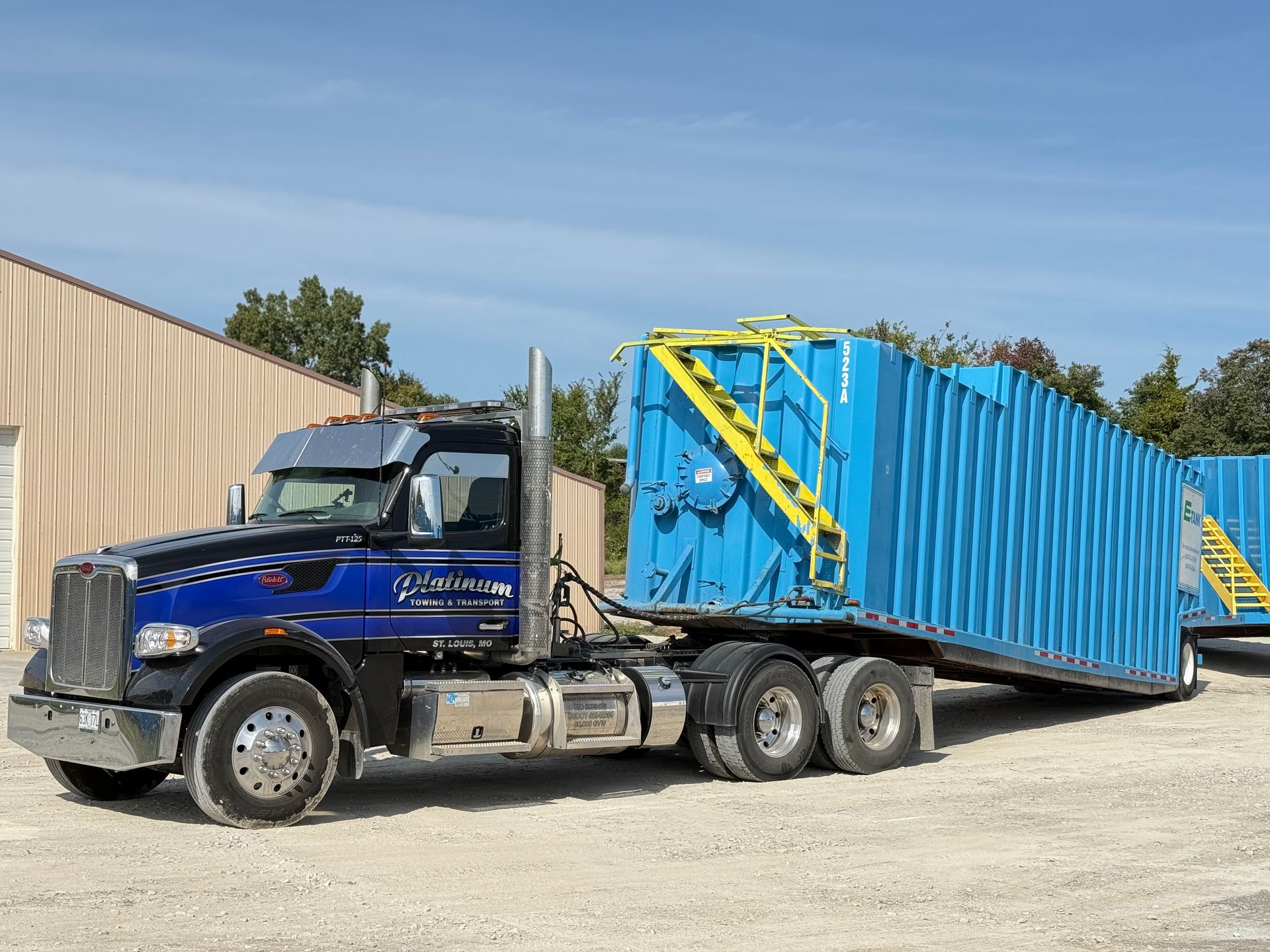 Blue semi-truck hauls two large blue rectangular tanks, possibly for industrial use, on a gravel lot under a blue sky.