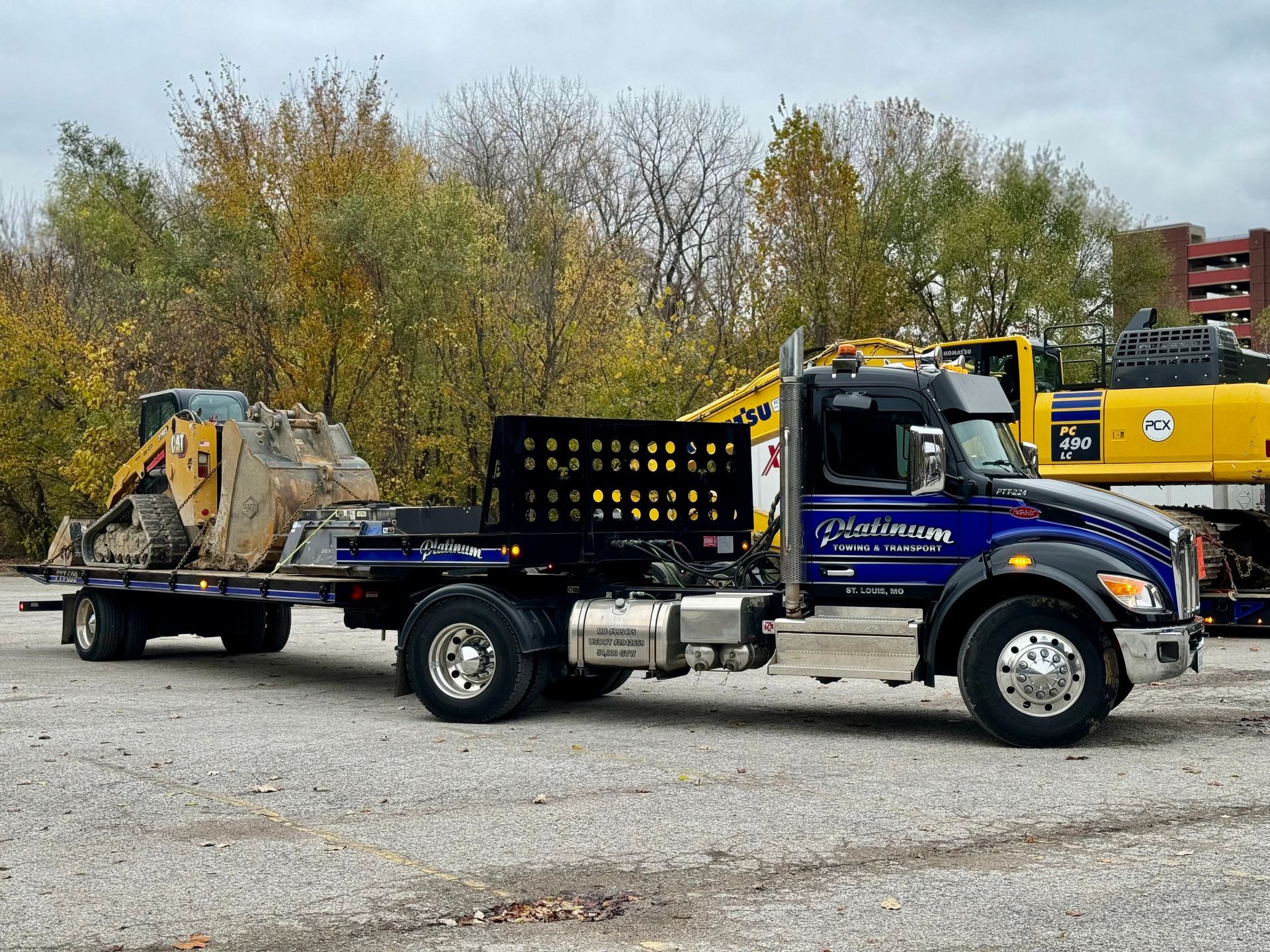 Blue truck with a trailer transporting a piece of construction equipment, parked on gravel.