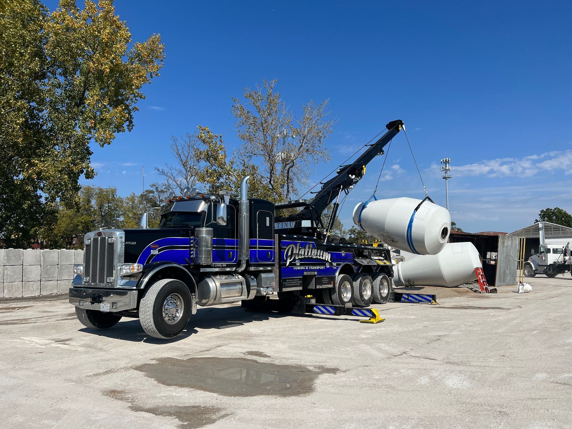 Blue tow truck lifting a white concrete mixer drum in a yard on a sunny day.