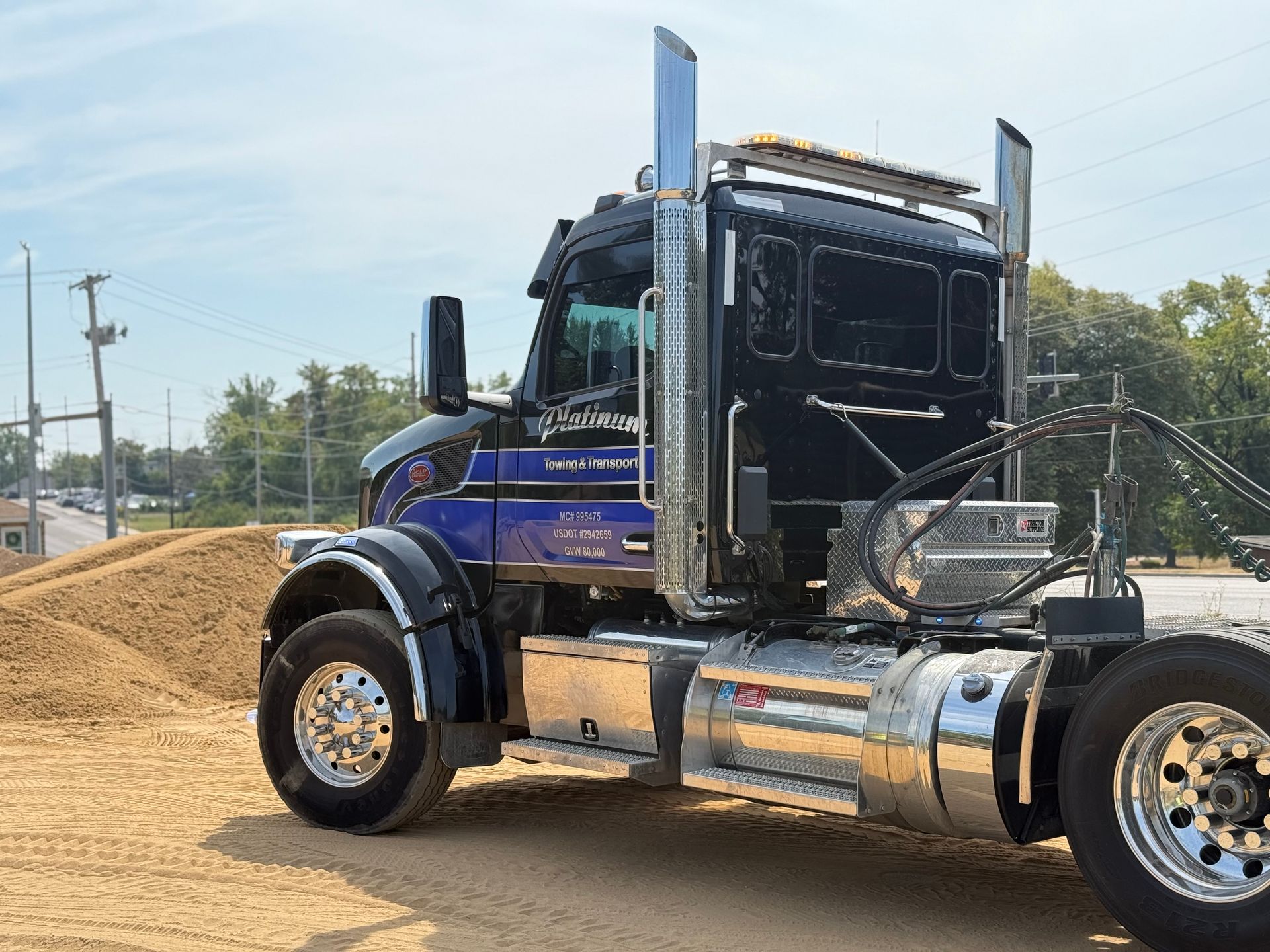 Black and blue semi-truck parked on a sandy lot with chrome details and tall exhaust stacks; sunny day.