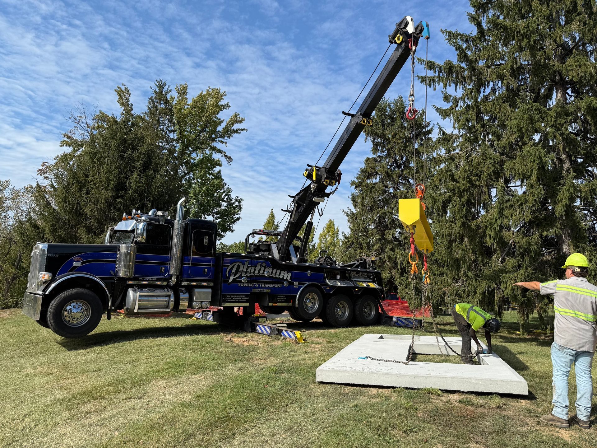 A blue tow truck crane lifting a yellow structure onto a concrete base, with a worker directing.
