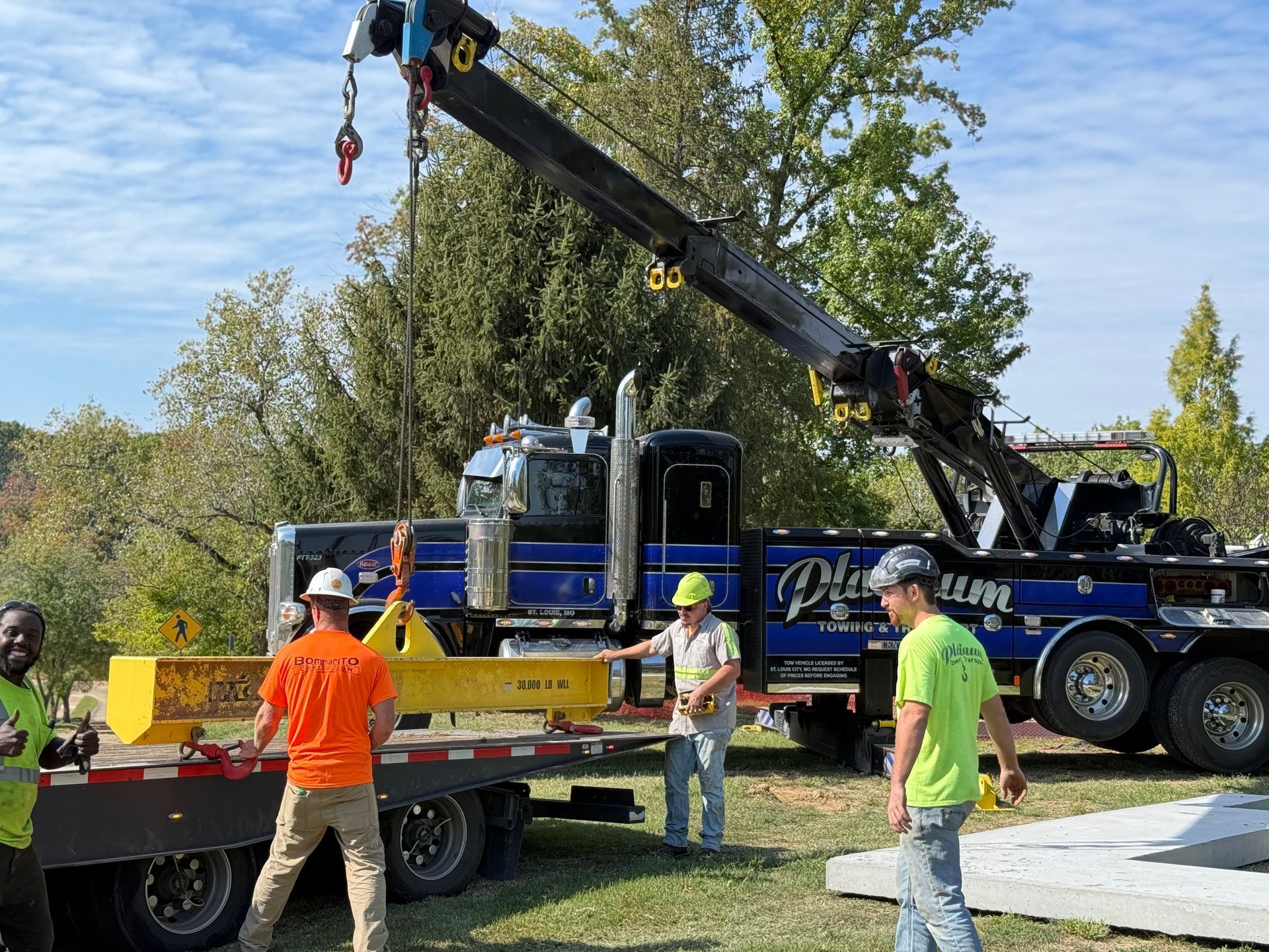 A tow truck with a boom lifting a yellow beam onto a flatbed trailer; several workers in safety vests nearby.