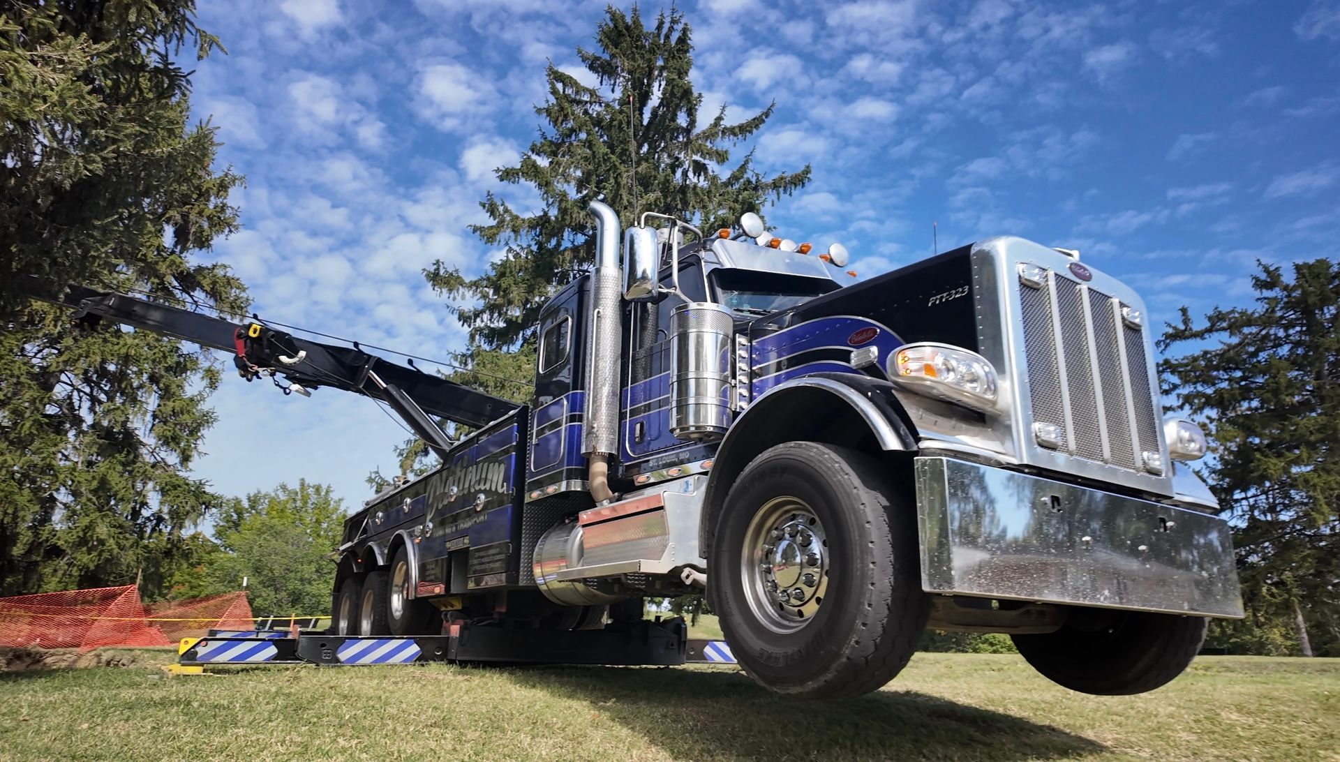 Blue tow truck on grass with boom extended, under a bright blue sky with trees.