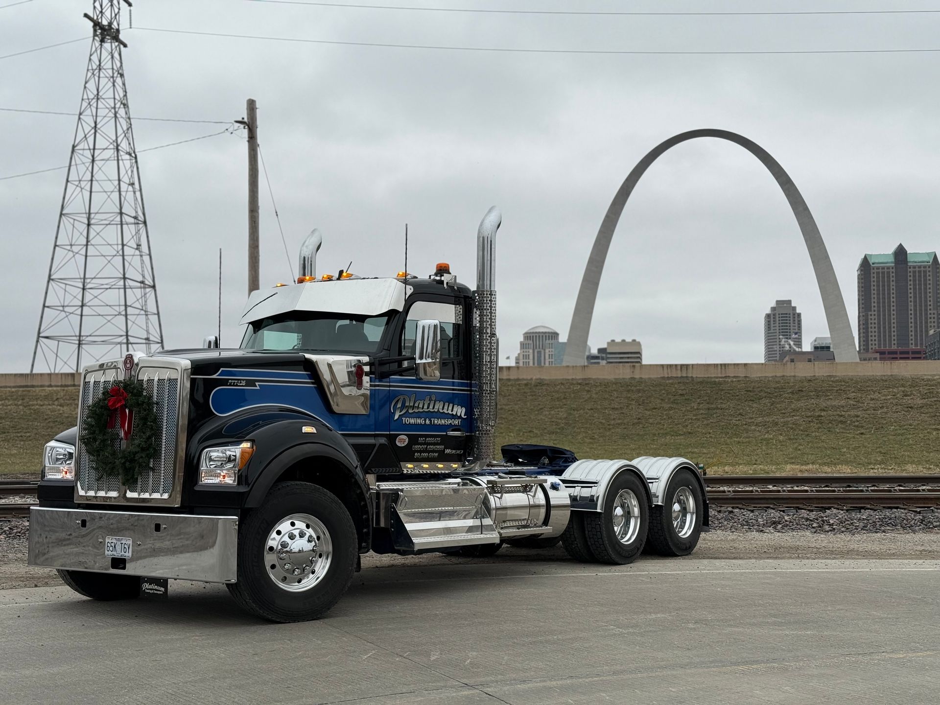Blue semi-truck with Christmas wreath in front of the Gateway Arch in St. Louis on an overcast day.