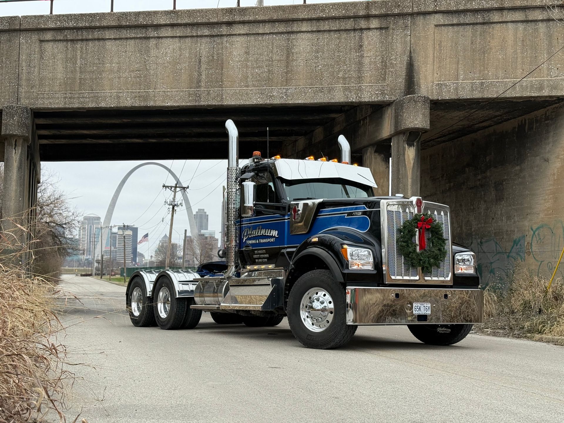 A blue semi-truck with a holiday wreath sits under a bridge, Gateway Arch in the background.