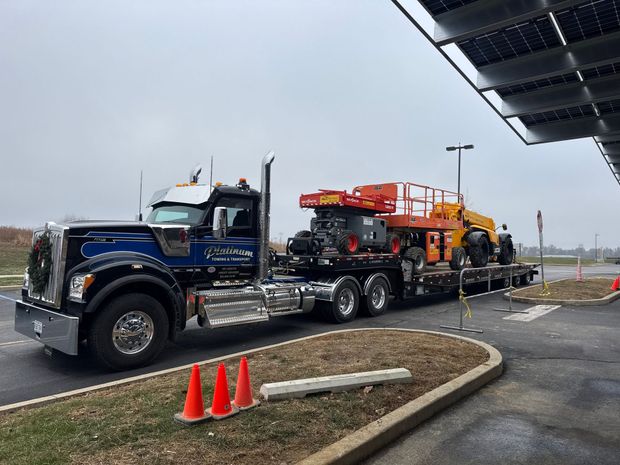 A semi-truck hauling heavy machinery, parked near a building with solar panels.
