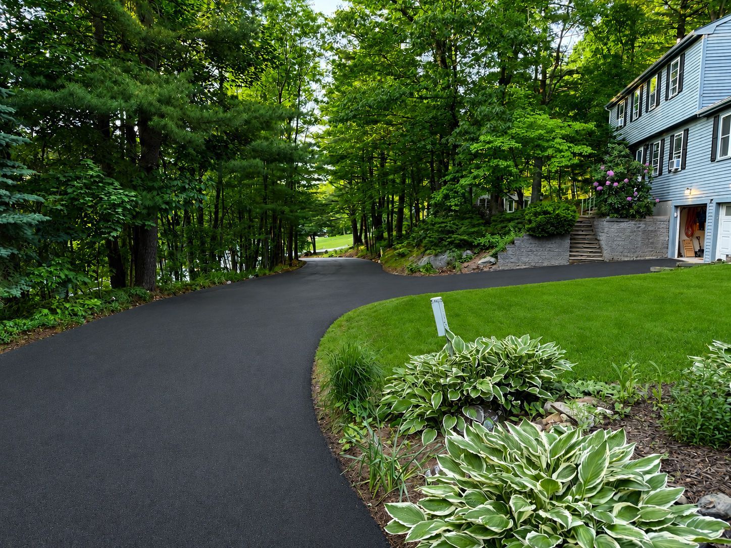 Curving black driveway beside lush green lawn and trees, with a house on the right