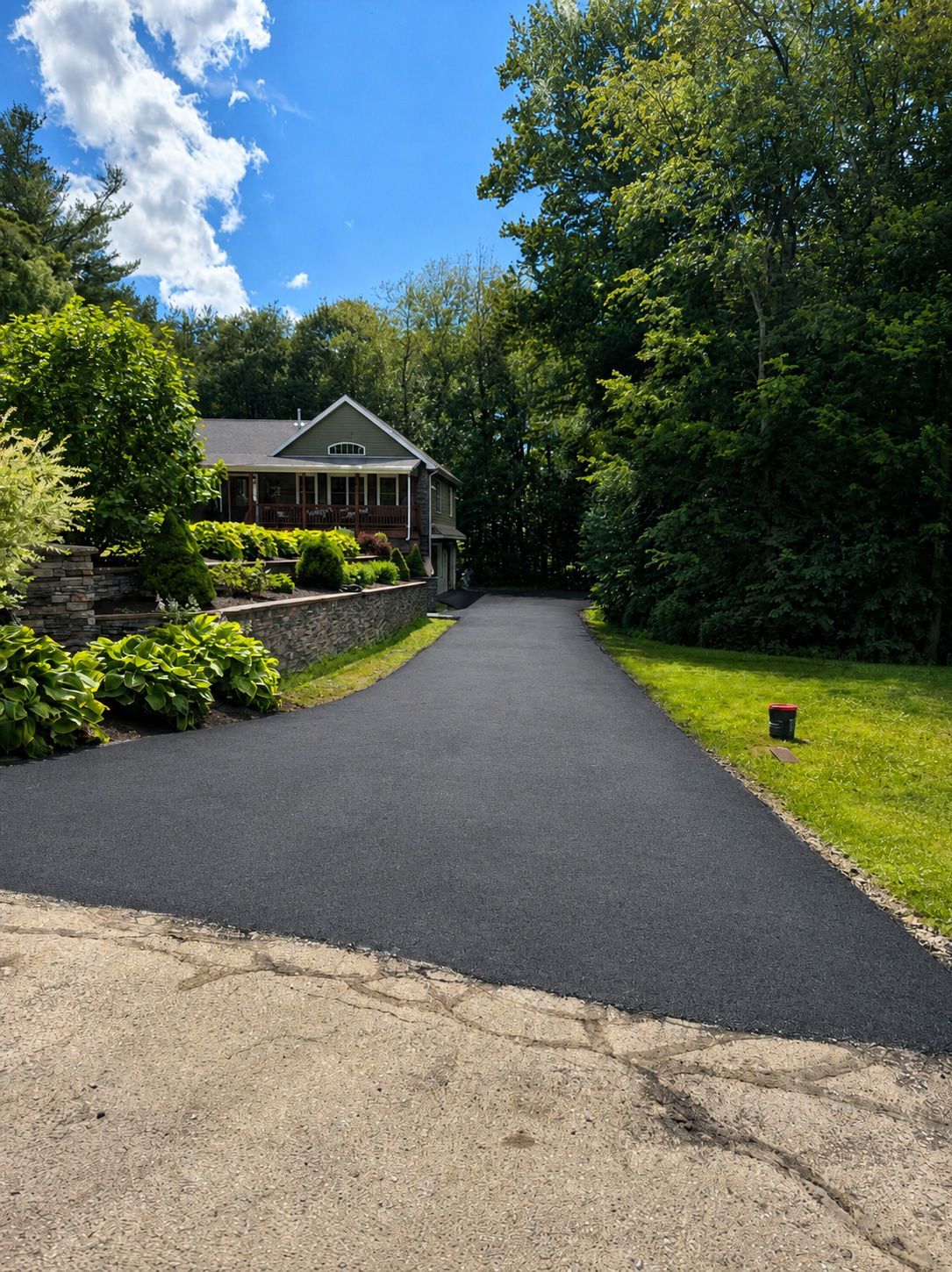 Fresh black driveway leading to a house surrounded by lush green trees and shrubs under a blue sky