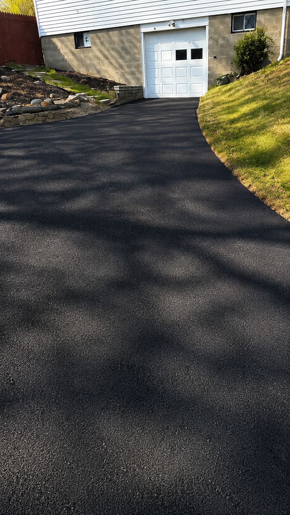 Freshly paved black driveway leading to a white garage beside a stone house and grassy slope