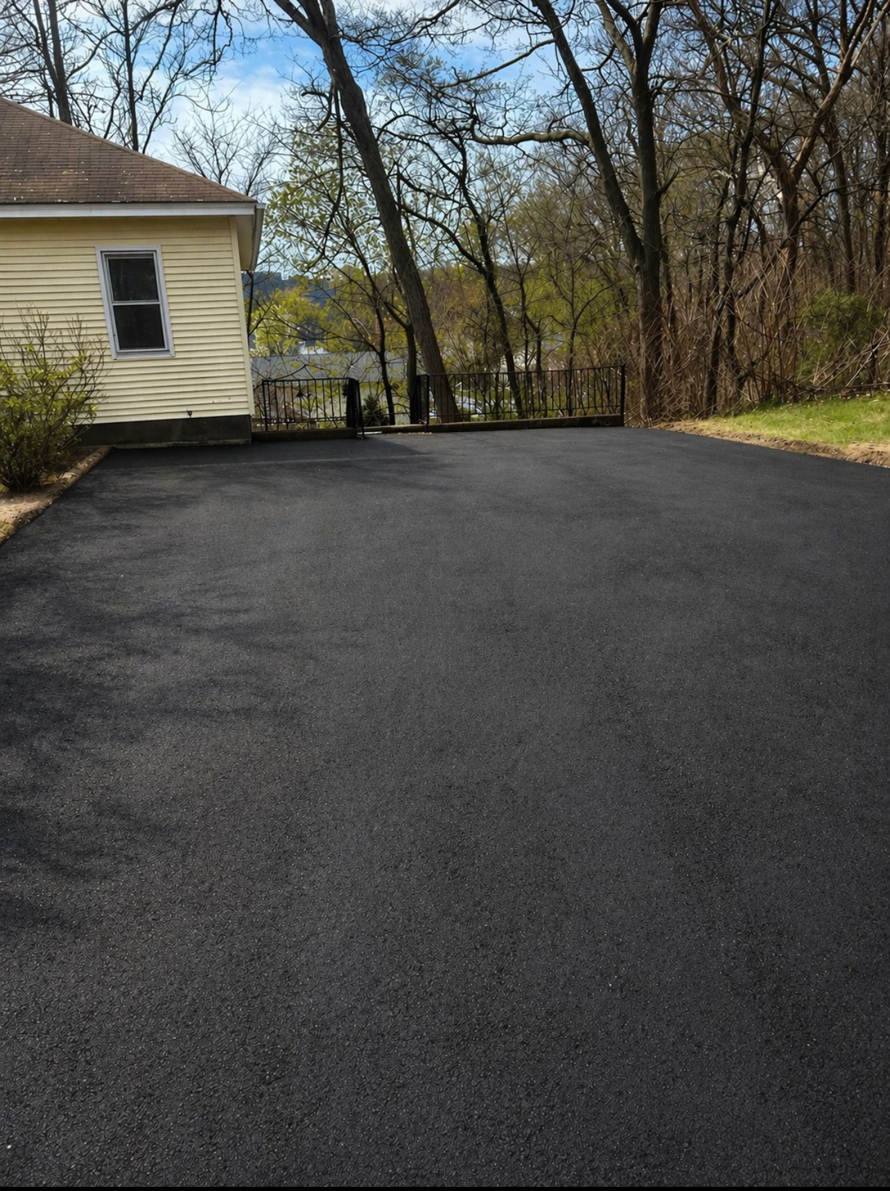 Fresh black asphalt driveway beside a yellow house, bordered by trees and woods