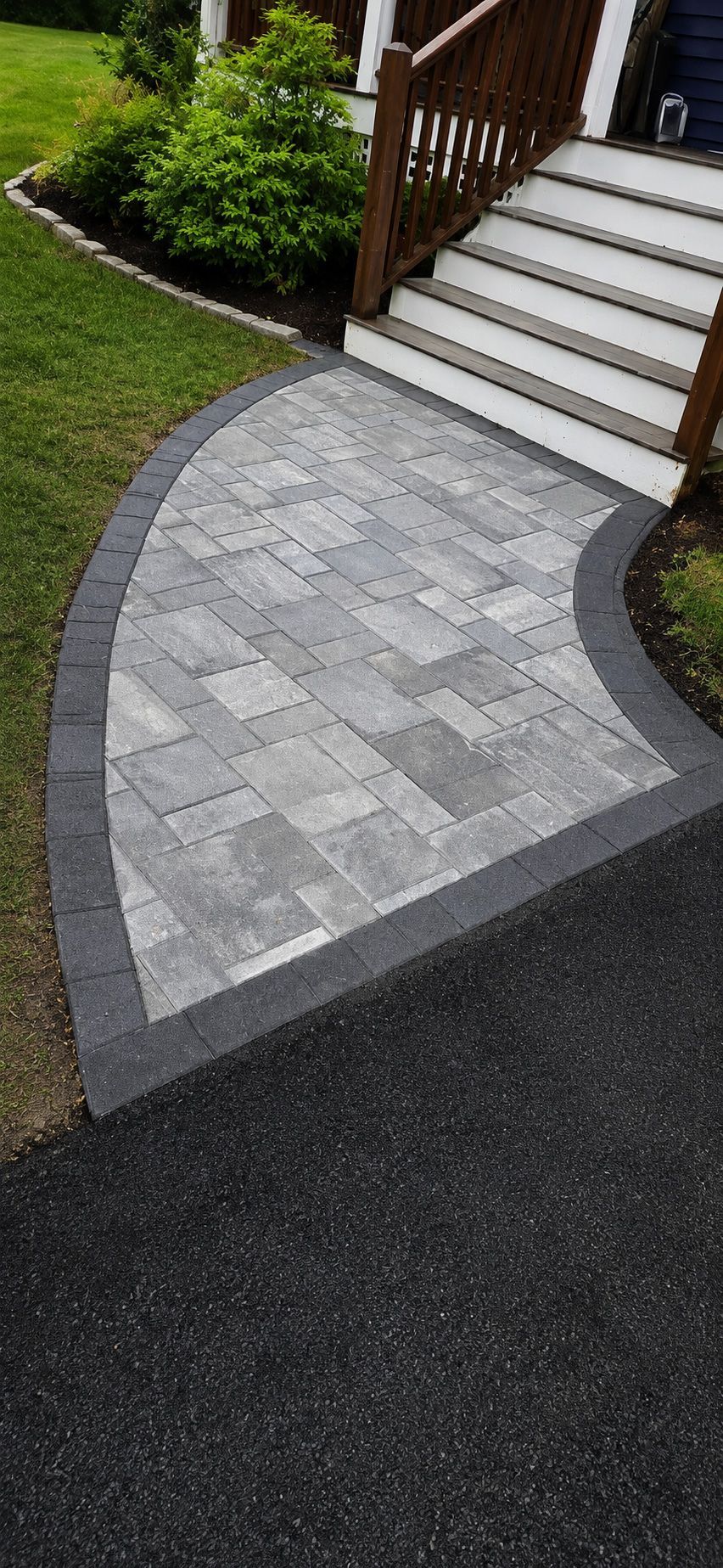 Curved gray paver walkway beside stairs, bordered by black asphalt and green lawn.