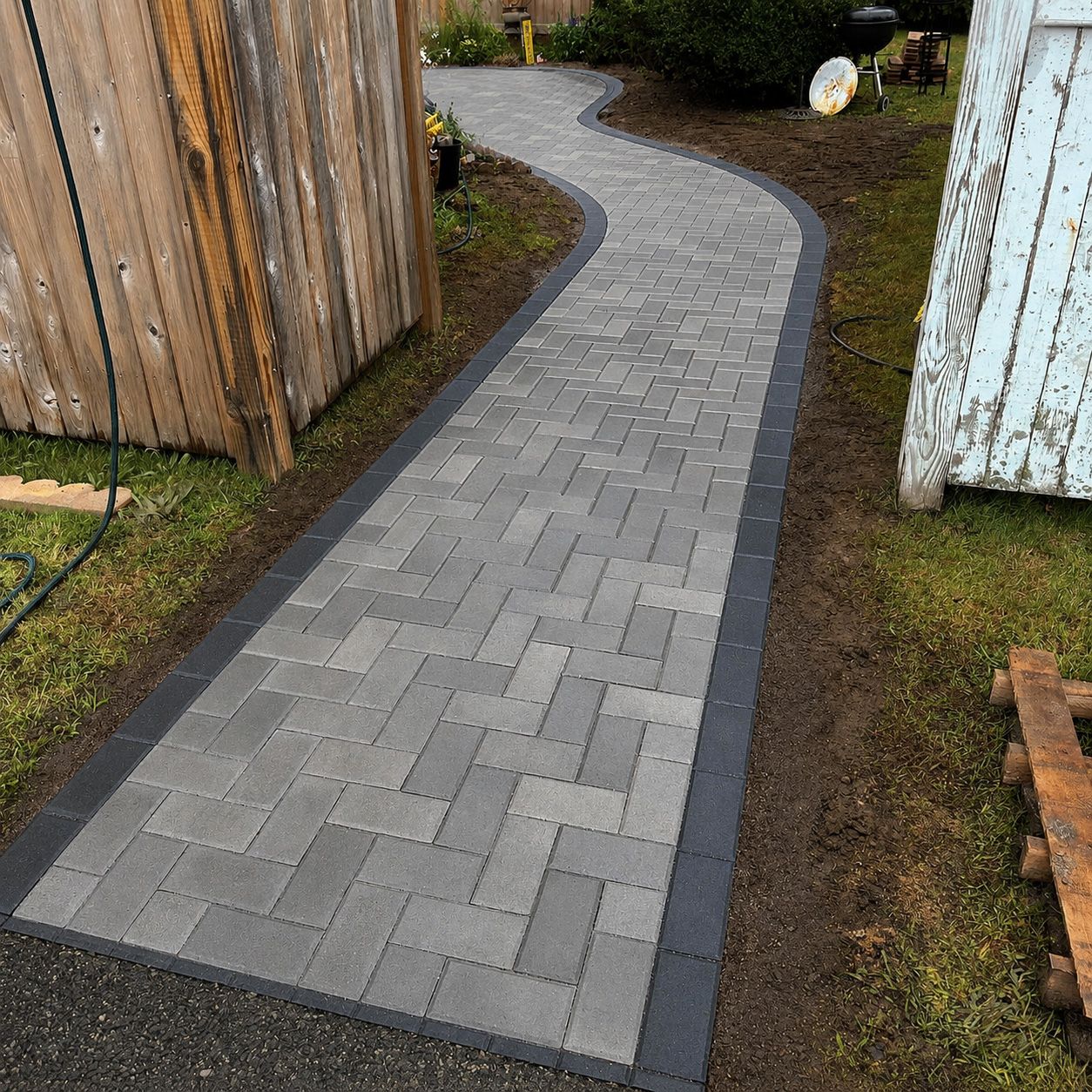 Curving gray paver walkway between wooden fence and shed, bordered by dark edging