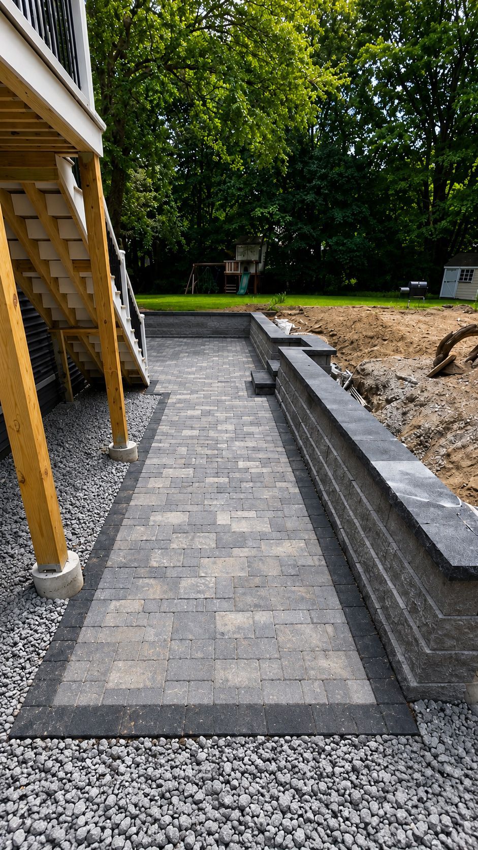 Gravel walkway beside a house, with gray pavers, retaining wall, and wooded yard beyond