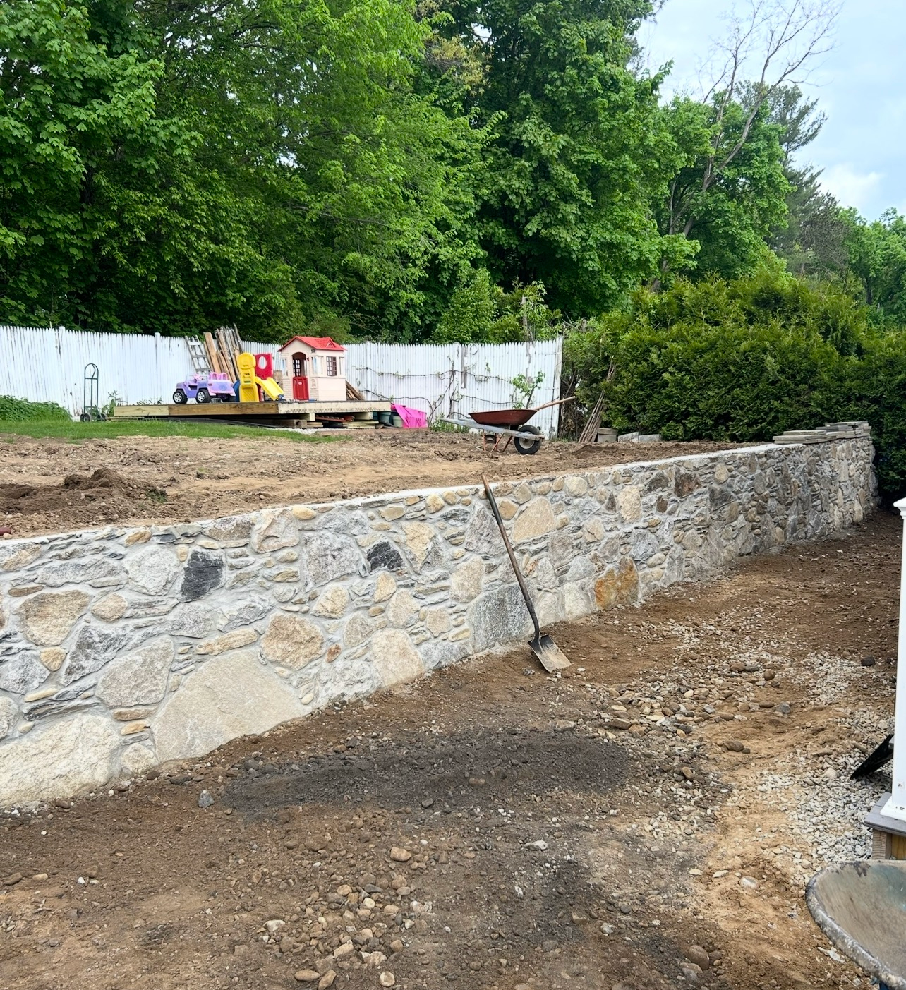 A newly built stone retaining wall runs across a backyard, with a small playhouse and a wheelbarrow visible behind it.
