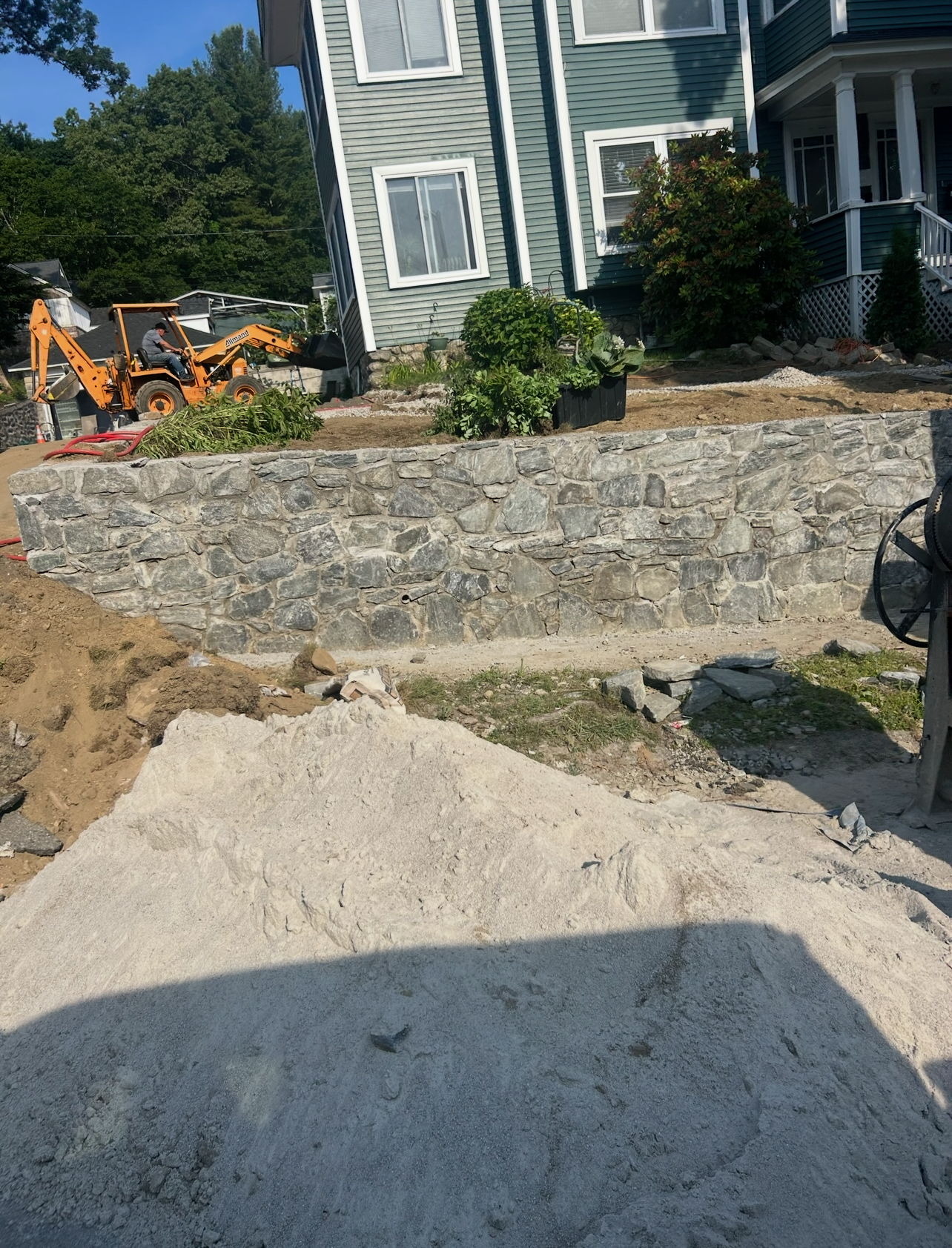 A large pile of gravel sits in the foreground before a stone retaining wall in front of a green residential house.