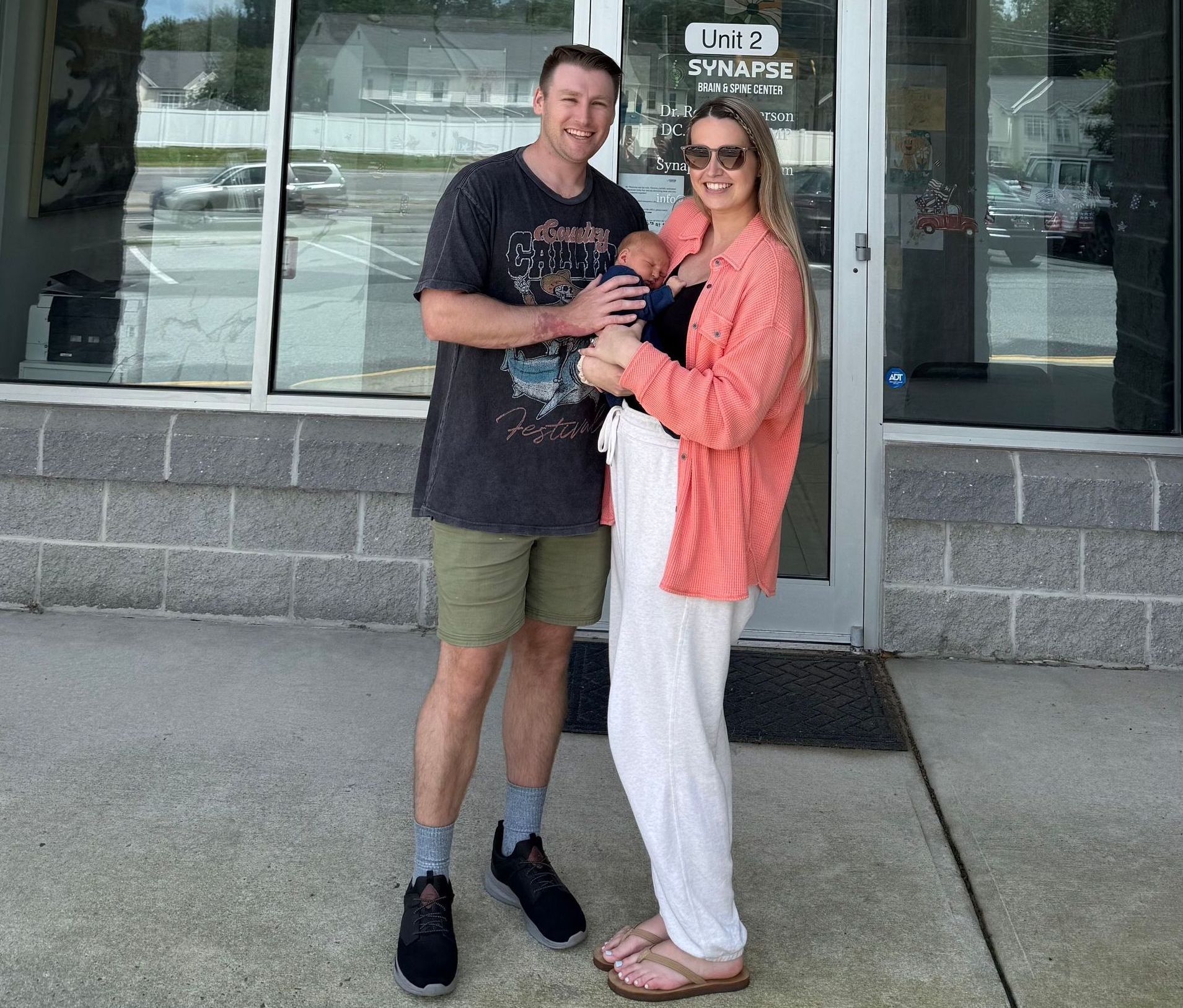 A couple stands outside a building, the woman holding a baby. The man wears a dark tee and shorts; the woman has a pink sweater and white pants.