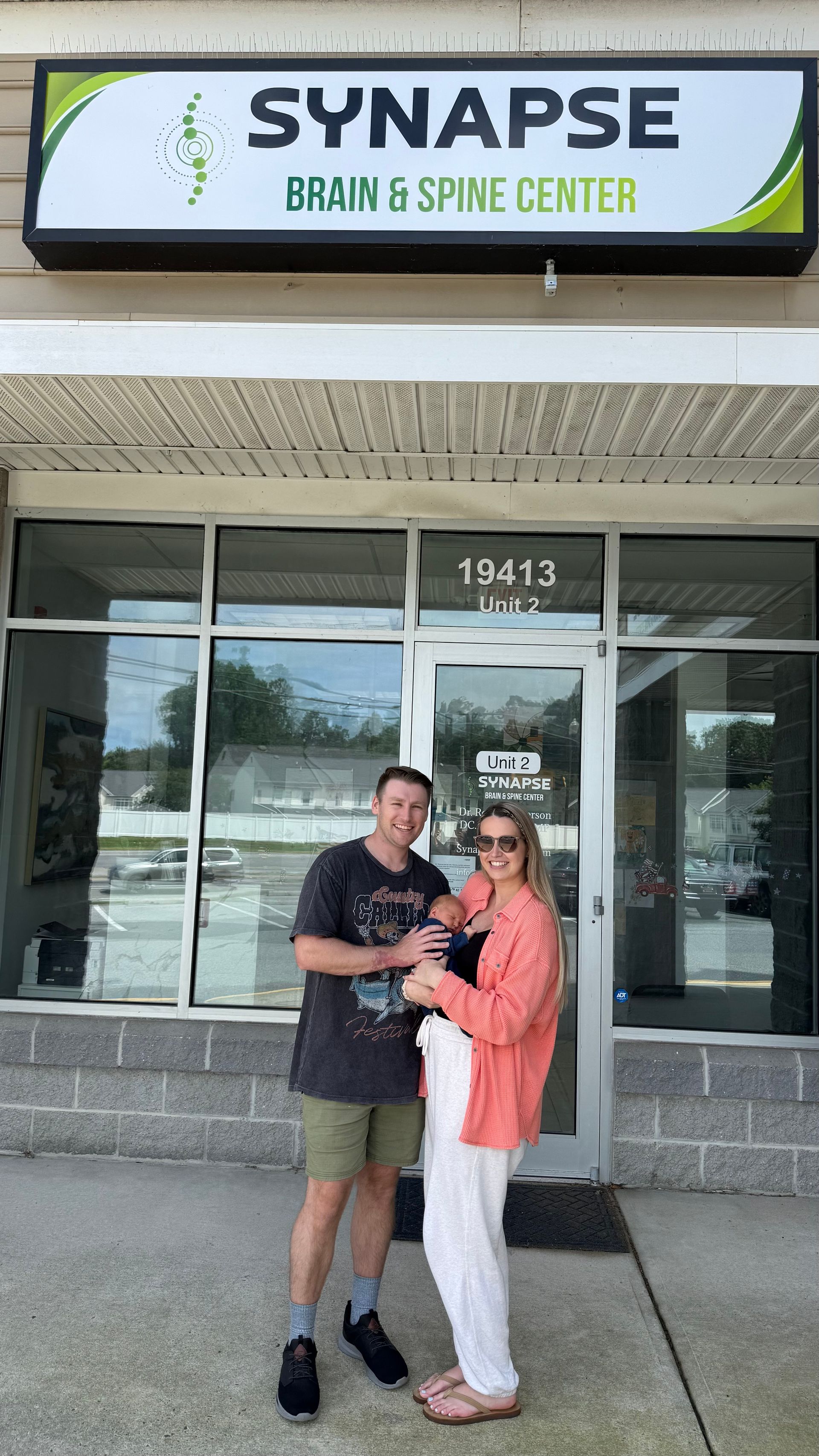 A couple holding a baby in front of the Synapse Brain & Spine Center building. They are smiling.